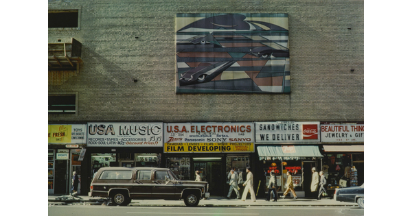 A color photograph of a brick building with colorful signs advertising products for different stores and a large work of art hanging above multiple stores.