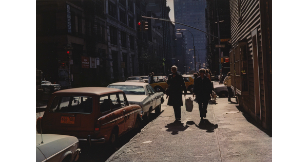 A color photograph of pedestrians on a city sidewalk next to parked cars, and cars passing through an intersection in the background.