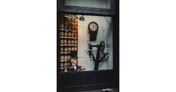 A color photograph of storefront window showing a shelf full of doll heads, a clock, and a piece of machinery.