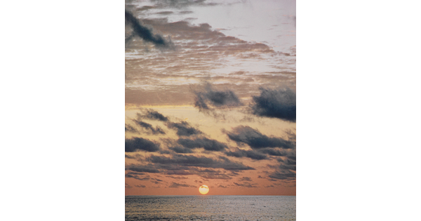 A color photograph of a sun just above an ocean horizon under wispy dark clouds.