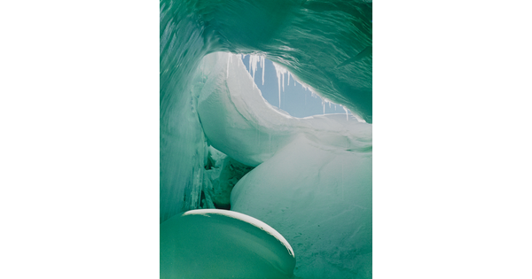 A color photograph from inside of a blue and green ice cave showing a sliver of blue sky.