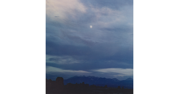 A color photograph of a mountain range below a full moon veiled behind thin dark clouds.