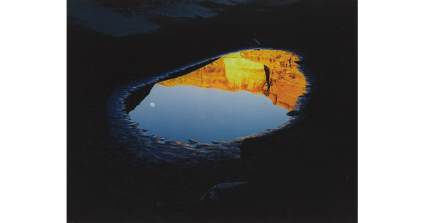 A color photograph of a pool of water reflecting a clear blue sky with a moon and orange mountain cliffs.