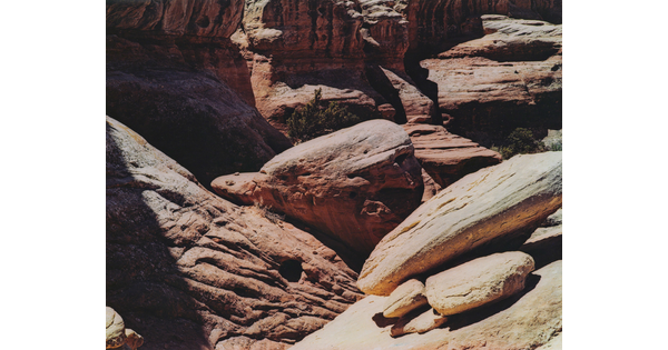 A color photograph of a red canyon with stark shadows and rocks balancing on a ledge in the foreground.