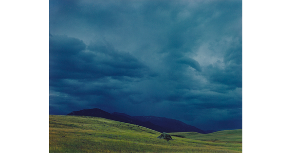 A color photograph of dark, heavy clouds above a mountain range beyond gentle green hills.