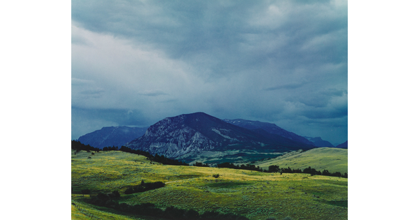 A color photograph of cloudy skies above a dark a mountain range beyond green fields in the foreground.