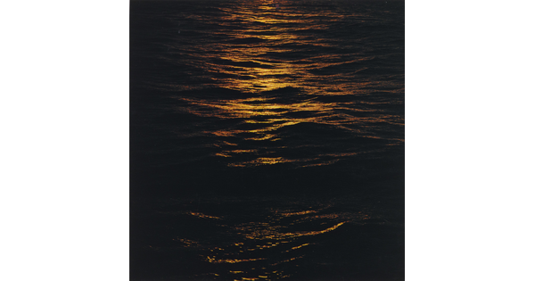 A color photograph of dark ocean waves at night reflecting orange light on the tops of the waves.