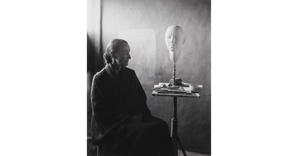 A black-and-white portrait photograph of a seated older White woman wearing a dark, floor length dress; a white face mask sits on a stand on a cluttered end table.