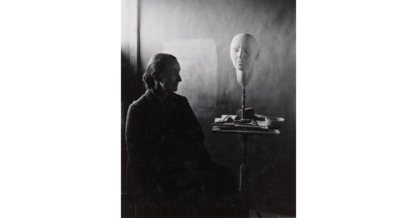 A black-and-white portrait photograph of a seated older White woman wearing a dark, floor length dress; a white face mask sits on a stand on a cluttered end table.