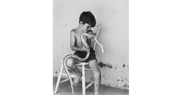 A black-and-white photograph of a shirtless young White boy sitting on a wooden chair holding a large snake.