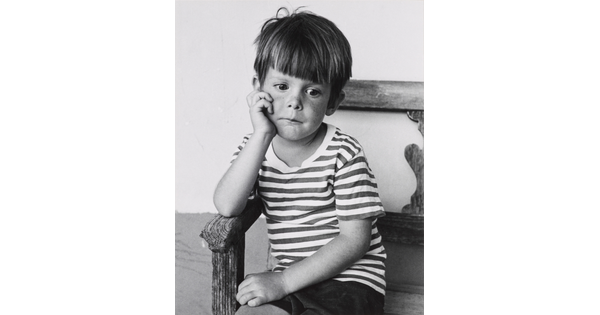 A black-and-white portrait photograph of a young White boy sitting in a wood chair resting his chin in his hand.