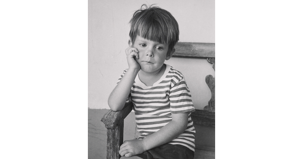 A black-and-white portrait photograph of a young White boy sitting in a wood chair resting his chin in his hand.