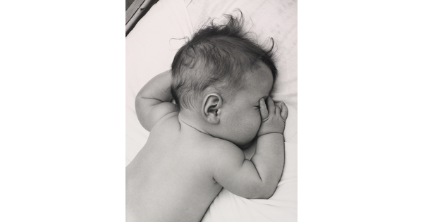 A black-and-white photograph of a White baby with dark hair lying on his stomach, thumb of one hand in his mouth, on white sheets.
