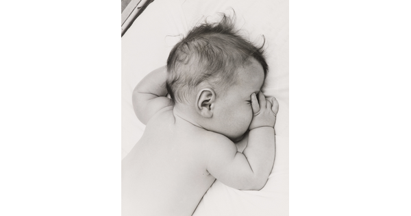 A black-and-white photograph of a White baby with dark hair lying on his stomach, thumb of one hand in his mouth, on white sheets.