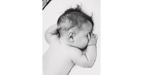 A black-and-white photograph of a White baby with dark hair lying on his stomach, thumb of one hand in his mouth, on white sheets.