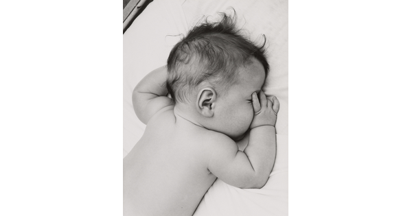 A black-and-white photograph of a White baby with dark hair lying on his stomach, thumb of one hand in his mouth, on white sheets.