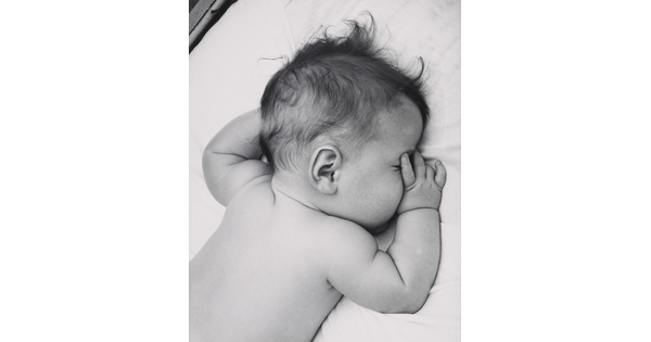 A black-and-white photograph of a White baby with dark hair lying on his stomach, thumb of one hand in his mouth, on white sheets.