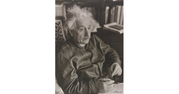 A black-and-white photograph of a seated older White man with shaggy hair and a mustache wearing a leather jacket; notepad on his lap.