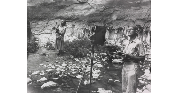 A black-and-white photograph of a White woman with a sketchbook and a White man with a camera next to a rock wall, both looking at something out of the frame.