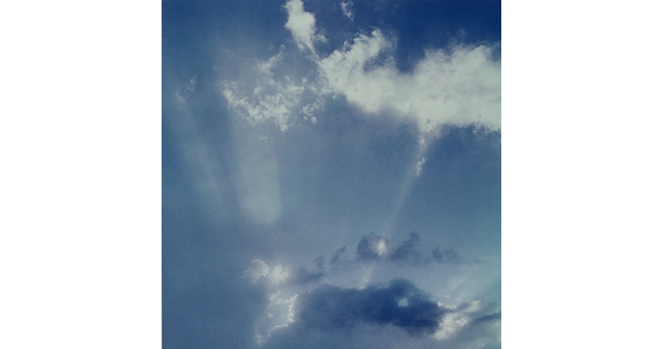 A color photograph of white clouds in a blue sky and sun rays that create white streaks.