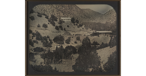 A black-and-white photograph of several buildings on a mountainside dotted with pine trees.