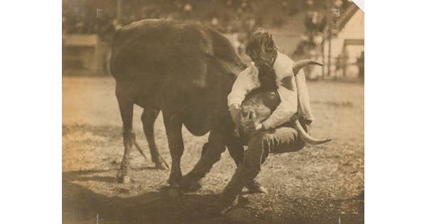 A sepia-toned photograph of a man with his arms wrapped around a bull's head from behind.