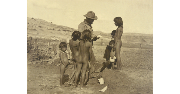 A sepia photograph of a group of mostly unclothed Indigenous children gathered around a White man in a barren landscape.