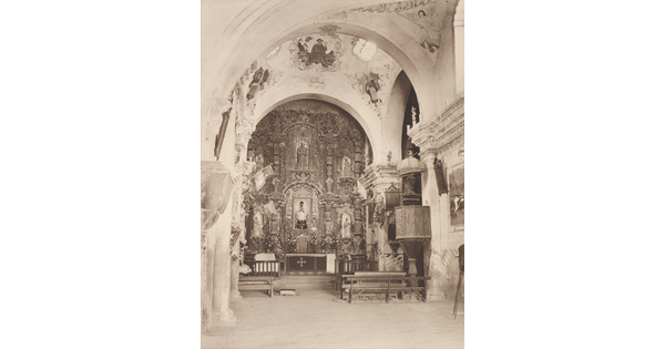 A black-and-white photograph of an ornate church interior showing arches and the altar.