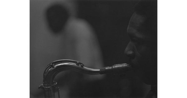 A black-and-white close-cropped photograph of a Black man in profile playing a saxophone.