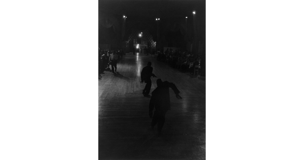 A dark black-and-white photograph of two male dancers in silhouette at the center of a dance floor.