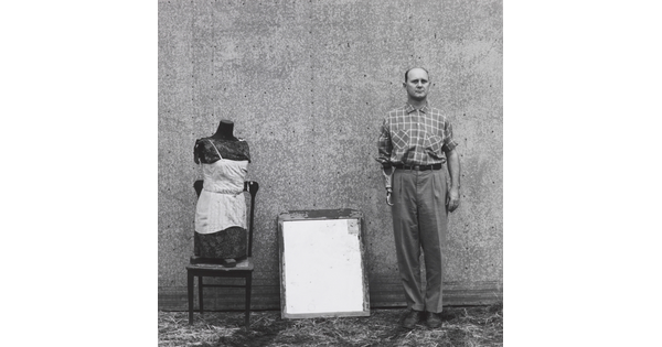 A black-and-white photograph of a mannequin bust on a wood chair, a mirror, and a balding White man with a prosthetic arm that ends in a hook lined up against a wall.