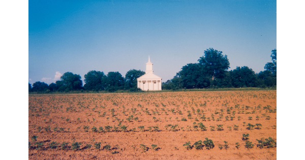A color photograph of a white wood church with a tall steeple and columns on the front sitting across an open field.