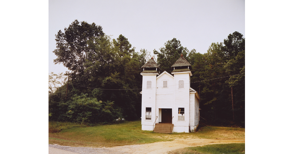 A color photograph of a white chapel situated in front of a wood.