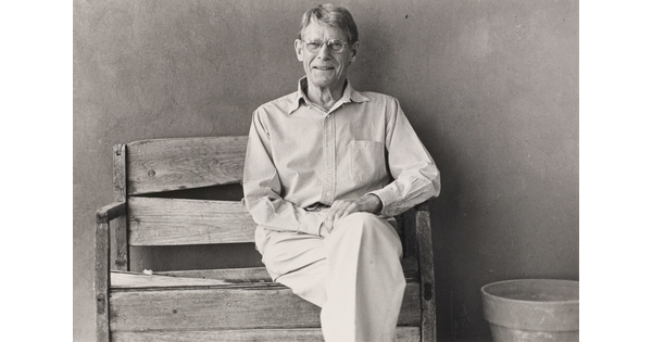 A black-and-white photograph of an older White man wearing glasses and a button-down shirt sitting on a wood bench.
