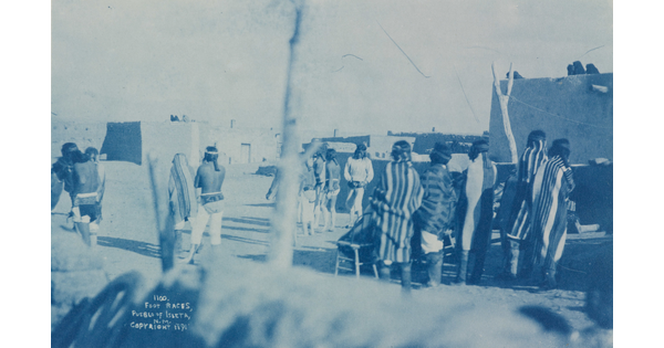 A blue-tinted photograph of Native American people, some wrapped in blankets, on a dirt road; several adobe buildings nearby.