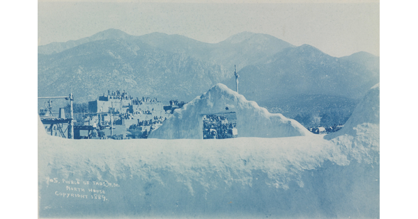A blue-tinted photograph of a multi-story pueblo at the base of mountains as seen from over an adobe wall.