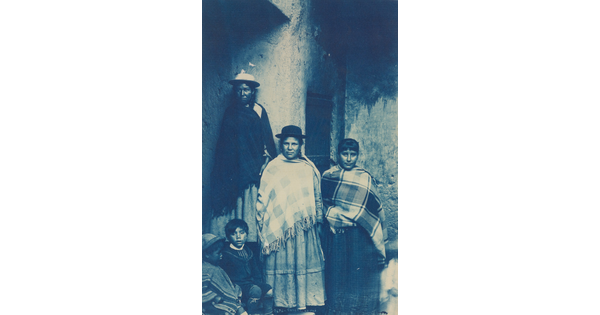 A blue-tinted photograph of women and children wearing ponchos standing next to a wall.