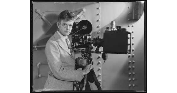 A black-and-white photograph of a White man wearing granny glasses and a light-colored suit standing next to an old movie camera.