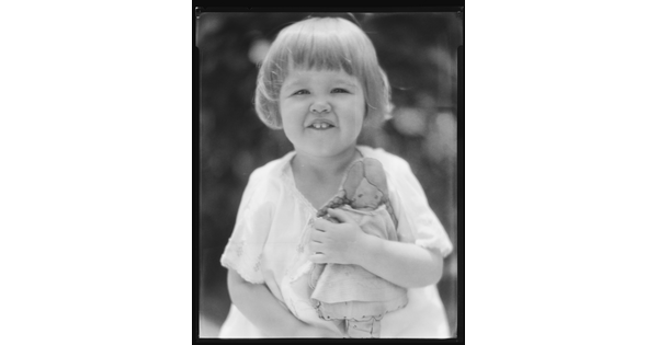 A black-and-white image of a young White child with short hair, showing her two front teeth, and holding a doll.