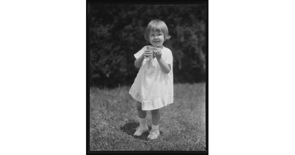 A black-and-white image of a young White child with short hair wearing a white dress and holding a mug as she stands in the grass.