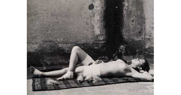 A black-and-white photograph of a young, topless White woman basking in sunlight next to sharp cacti.