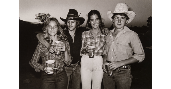 A black-and-white photograph of White teenagers, two girls and two boys with their arms around each other, holding Coca-Cola cups and wearing western clothing.