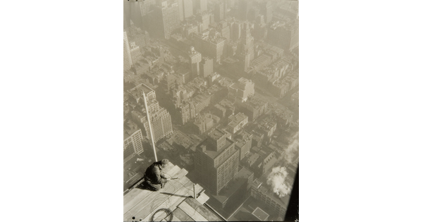 A black-and-white photograph taken from above of a man kneeling precariously on the edge of a construction platform suspended high over a city skyline.