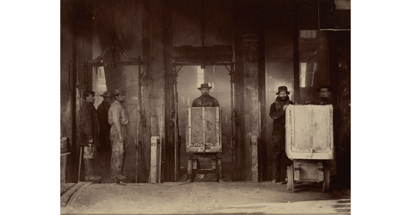 A sepia-toned photograph of men pushing minecarts towards the viewer in an industrial setting with beams of light back-lighting them.