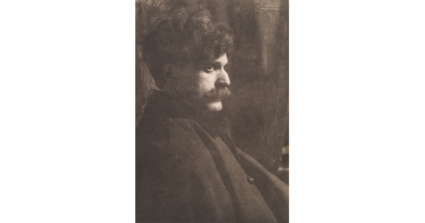 A black-and-white photograph of a White man with lots of dark, curly hair and a mustache sitting in profile.