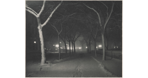 A black-and-white photograph of a snowy street lined with bare trees at night, streetlamps glowing in the distance.