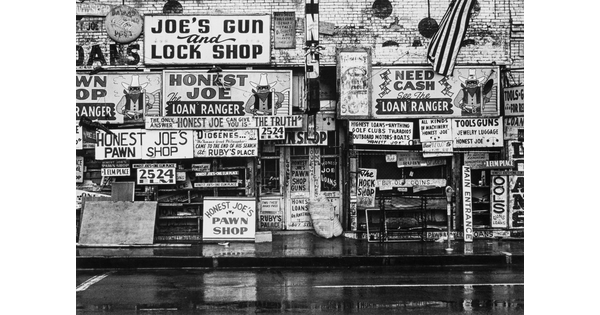 A black-and-white photograph of the outside of a pawn shop covered in signs advertising "Honest Joe" and the "Loan Ranger."