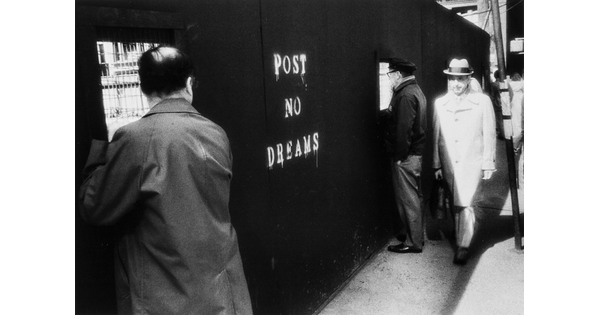 A black-and-white photograph of men looking through the windows of a construction wall; the wall has "Post no dreams" painted on it.