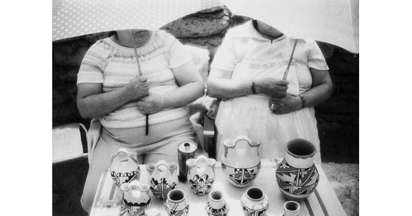 A black-and-white photograph of two women, faces hidden by umbrellas they are holding, at a table filled with pottery decorated with Native American designs.