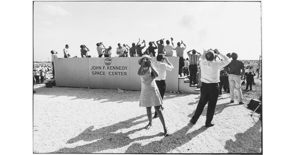 A black-and-white photograph of crowd of people looking up into the sky from a viewing stand on a beach with a sign that reads, "John F. Kennedy Space Center."